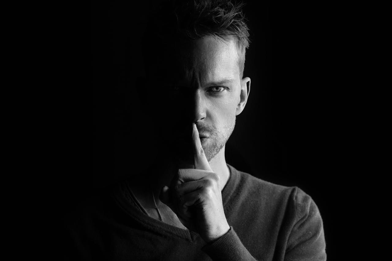 Black and white portrait of a man gesturing for silence, highlighting mystery.